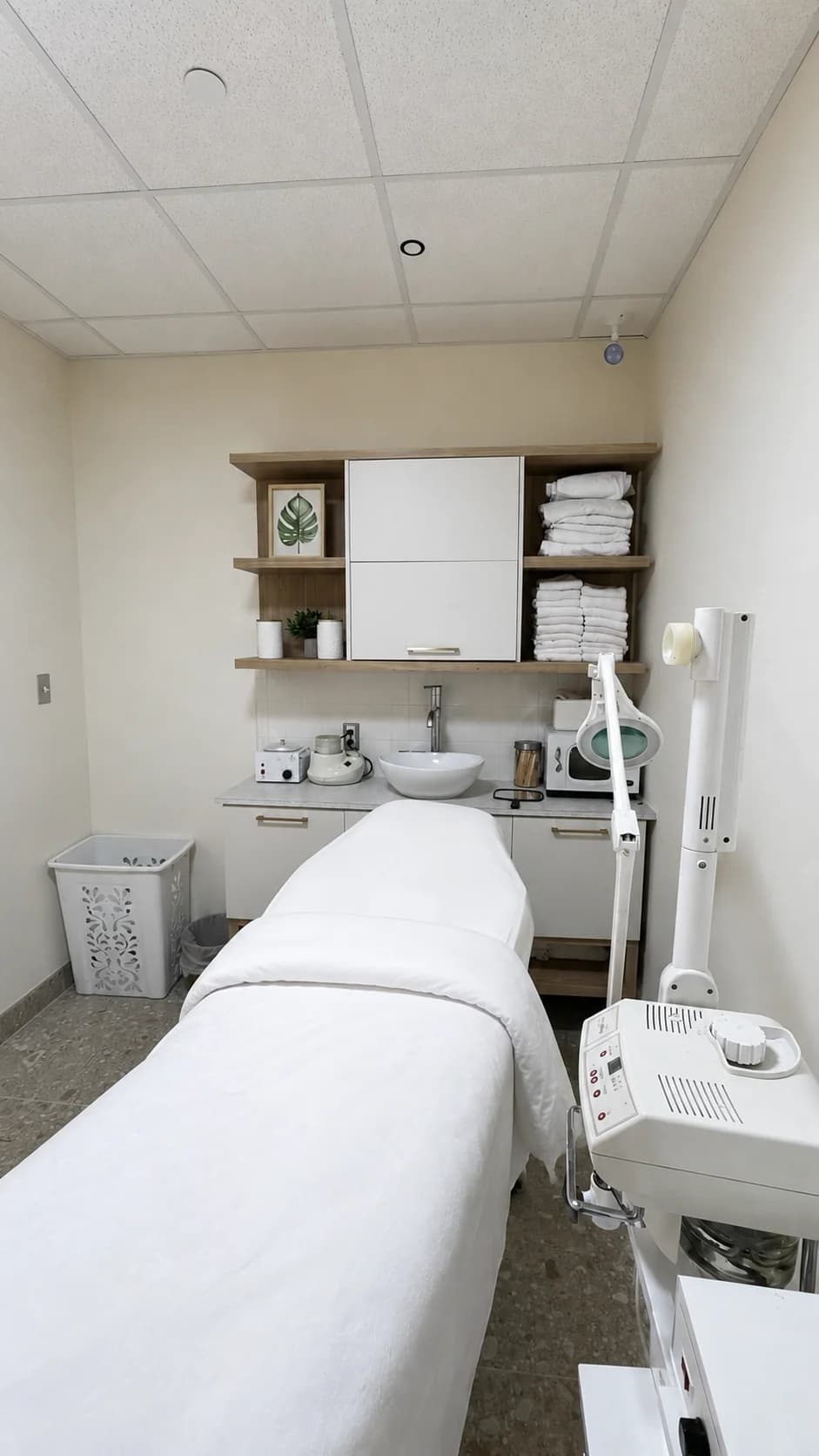 Salon treatment room with a white-covered bed, storage shelves, and treatment equipment